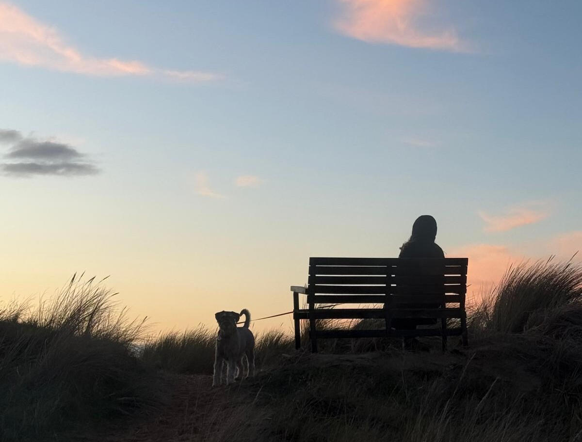Woman seated on a bench beside her dog during a quiet sunset, open sky in the background.